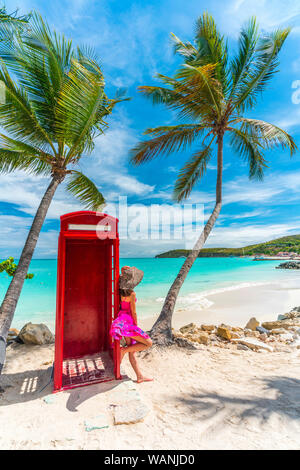 Woman in elegant dress and hat looking at sea from the old red phone box, Siboney Beach, Dickenson Bay, Antigua, Caribbean Stock Photo