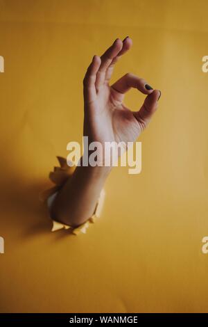 Vertical shot of a person's hand breaking through a yellow paper wall showing an 'OK' sign Stock Photo