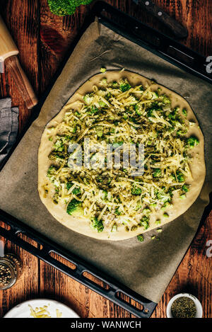Homemade pizza with broccoli, pesto sauce, spices and cheese in a baking tray ready for the oven. View from above Stock Photo
