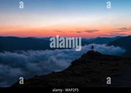 A hiker stands on a high, rocky ridge and looks out towards the Never Summer Range as fog fills the canyon below at sunset in Rocky Mountain National Stock Photo