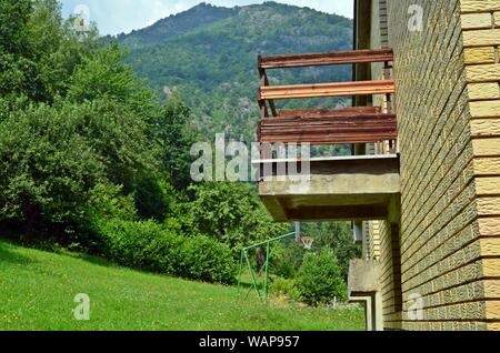 A wooden balcony of a yellow brick mountain village house with a basketball outdoor court and a mountain in the background, a forest on the left side Stock Photo