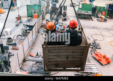 Two men are moving on an industrial crane in the factory floor. Bloggers and journalists are filming a story about the production of an engineering pl Stock Photo