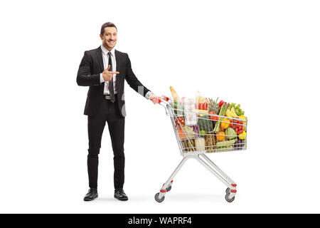 Full length portrait of a young man in a suit standing with a shopping cart full of food and pointing isolated on white background Stock Photo