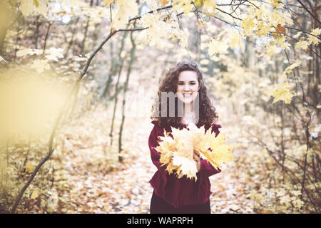 Beautiful girl with curly dark hair in a maroon top in an autumn park holding maple leaves and smiling at the camera Stock Photo
