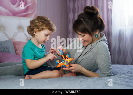 Stylish mother with a charming little son spend time together in the bedroom. Mom with baby play toys on the parent's bed. Having fun together. Intere Stock Photo