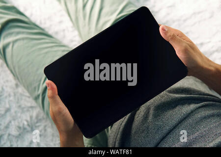 Lower half of a sitting man on the bed looking down to empty black screen of an up-to-date tablet. Young man at home looks at tablet screen. Switched Stock Photo