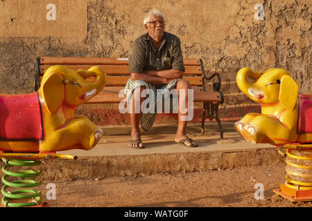 On a late afternoon, an elderly man relaxes on a bench in a children's playground, framed by two rocking elephants; in Walkeshwar area, Mumbai, India Stock Photo