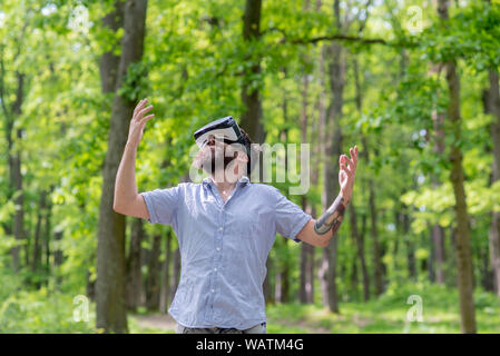 Happy man using VR glasses in forest or park. Hipster in blue shirt enjoying 3D view in modern device. Bearded man wearing virtual reality goggles Stock Photo