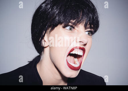 Screaming female with crazy look isolated on gray background, emotions concept. Bad tempered businesswoman in black jacket wearing dark wig expressing Stock Photo