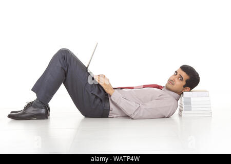 Businessman lying against stack of books and using laptop Stock Photo