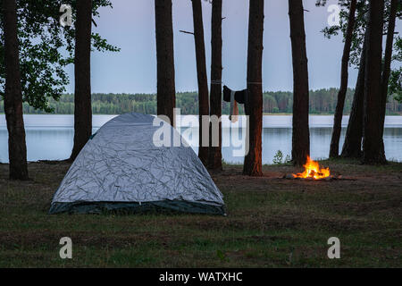 View of the campsite by the lake at twilight. Tent and burning fire place by the water surrounded by trees in the forest. Spending the night in wilder Stock Photo