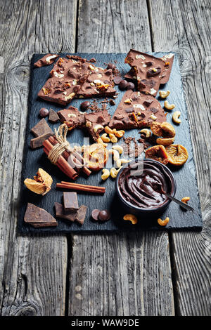 homemade sweet chocolate bar with dried figs and cashew filling on pieces on a black slate tray with ingredients, vertical view from above Stock Photo