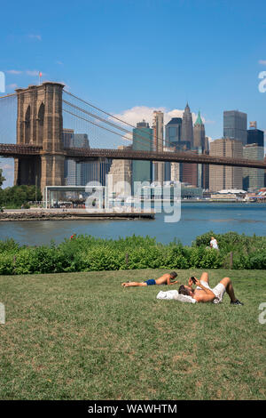 Brooklyn summer, view of men sunbathing in Main Street Park, Brooklyn, with the Brooklyn Bridge and Lower Manhattan skyline in the distance, NYC, USA Stock Photo