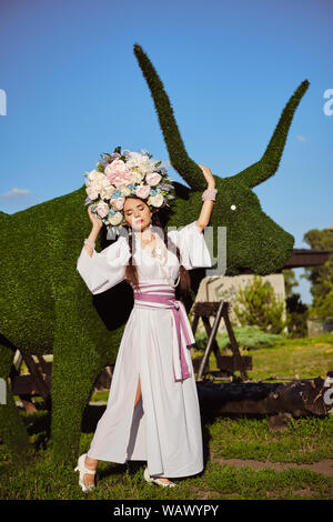 Brunette girl in a white ukrainian authentic national costume and a wreath of flowers is posing against a green ox carved from a bush. Stock Photo