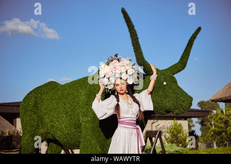 Brunette girl in a white ukrainian authentic national costume and a wreath of flowers is posing against a green ox carved from a bush. Stock Photo