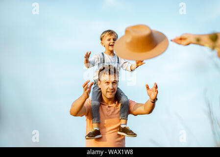 Portrait of a happy father with young son riding on the shoulders, playign with hat on the blue sky background outdoors. Concept of a happy family on Stock Photo