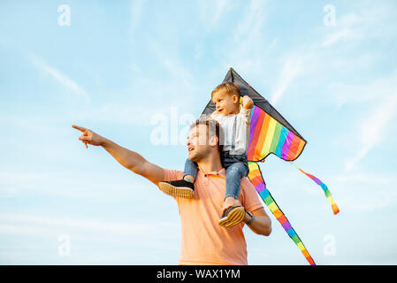 Portrait of a happy father and young son on the shoulders with colorful air kite on the blue sky background. Concept of a happy family and summer acti Stock Photo