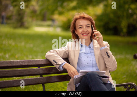 one smiling woman, looking at camera, while talking over phone. sitting in park on a bench. Stock Photo
