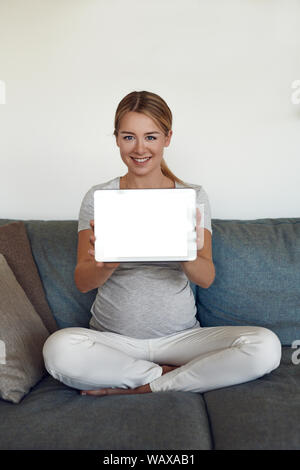 Smiling attractive vivacious young pregnant woman sitting cross legged on a sofa at home holding up a tablet with blank screen and copy space Stock Photo