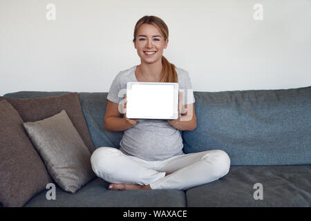 Smiling attractive vivacious young pregnant woman sitting cross legged on a sofa at home holding up a tablet with blank screen and copy space Stock Photo