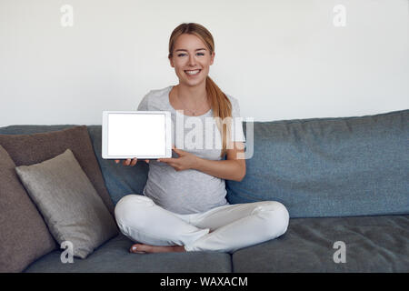 Smiling attractive vivacious young pregnant woman sitting cross legged on a sofa at home holding up a tablet with blank screen and copy space Stock Photo