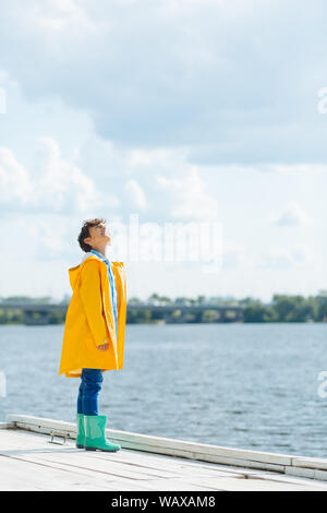 Cheerful boy wearing yellow raincoat looking at cloudy sky Stock Photo