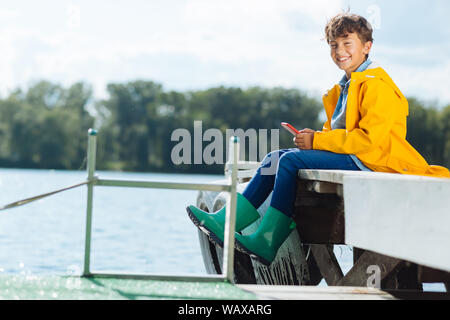 Boy wearing green rain boots smiling while sitting near river Stock Photo