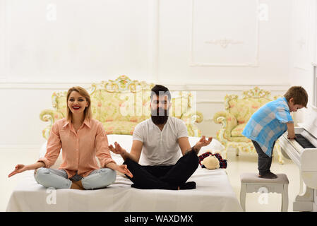 Young parents meditating, while their little son playing piano, annoying parents. Kid annoyed parents with noise. Mom and dad try to keep calm. Medita Stock Photo