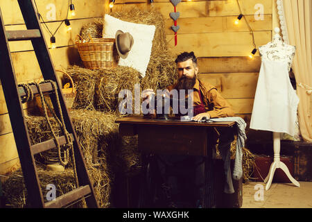 Handsome man tailor or dressmaker with beard and moustache sews clothes on vintage sewing machine in rustic workshop Stock Photo