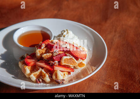 Crispy waffle with fresh strawberries baked walnuts whipping cream and honey in white plate on wood table under sunlight. Stock Photo