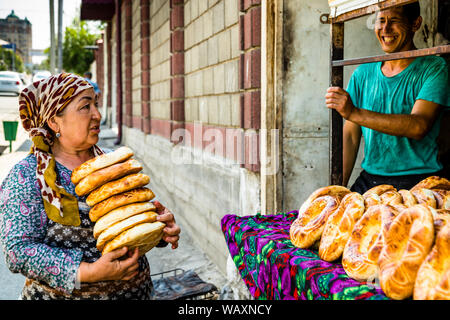 A woman carries a tall stack of fresh flatbreads, Tajik Non (Naan), in her arms as the laughing baker looks out from the sales window behind her, Osh, Kyrgyzstan. Stock Photo