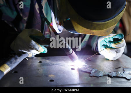 Professional female welder welding metal parts with electric torch wearing protective mask and gloves in warehouse.Industrial workshop worker using sp Stock Photo