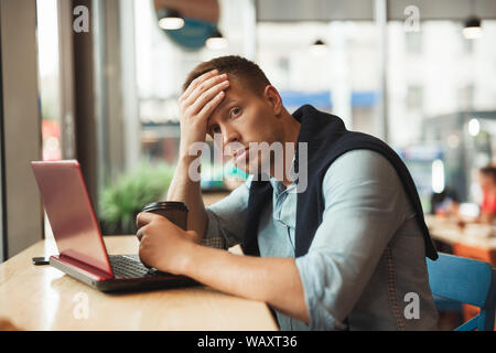 young handsome man works in his laptop in cafe eating croissant and drinking hot coffee for lunch feeling tired Stock Photo