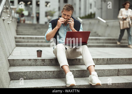 young handsome business man having his lunch on the go sitting on the stairs working in laptop having conversation in the city Stock Photo
