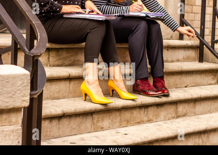 Two caucasian females holding a business meeting outdoors while smoking a cigarette, hands, legs and feet with extravagant shoe wear are to be seen Stock Photo