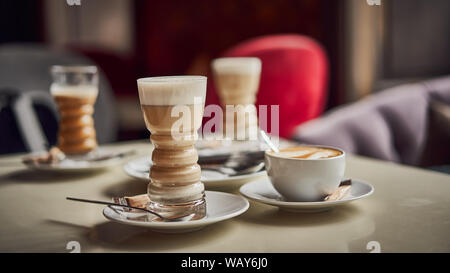 Latte glass with layered latte, cappuccino or mocha with foam on table in cafe with milk saucer and spoon. Three cups of coffee. Food and drink toning Stock Photo