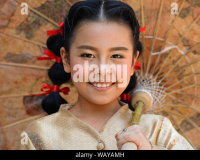 Dressed-up Thai pre-teen girl with red hair ribbons poses for the camera under a traditional Chinese oil-paper parasol. Stock Photo