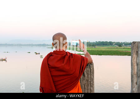 MANDALAY, MYANMAR - 03 DECEMBER, 2018: Horizontal picture of burmese monk taking pictures of the river from U Bein Bridge during sunset in Mandalay, M Stock Photo