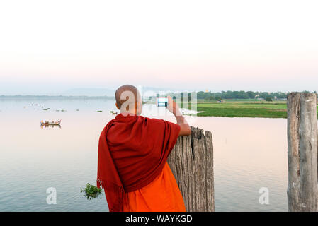 MANDALAY, MYANMAR - 03 DECEMBER, 2018: Horizontal picture of buddhist monk taking pictures of the river from U Bein Bridge during sunset in Mandalay, Stock Photo