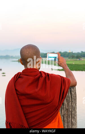 MANDALAY, MYANMAR - 03 DECEMBER, 2018: Vertical picture of buddhist monk taking pictures of the river from U Bein Bridge during sunset in Mandalay, My Stock Photo