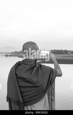 MANDALAY, MYANMAR - 03 DECEMBER, 2018: Black and white picture of buddhist monk taking pictures of the river from U Bein Bridge during sunset in Manda Stock Photo