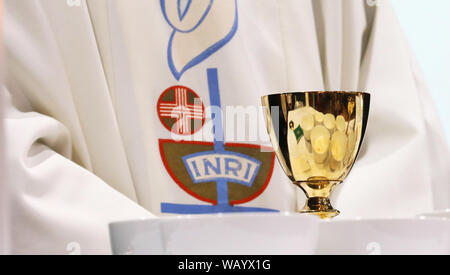 Close up of a priest wearing a white gown with the word INRI embroidered on to it. A single brass chalice with reflected bowls containing the host Stock Photo