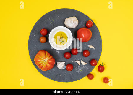 some tomatoes, garlic cloves and a bowl with olive oil on a black stone plate and a yellow background on a white surface Stock Photo
