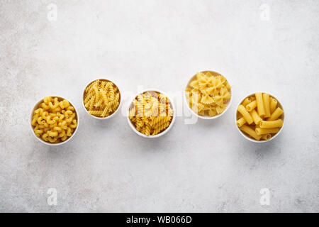 Various types of dry pasta in bowls on light gray background. Fusilli, farfalle, cavatappi, girandole and tortiglioni. Flat lay. Copy space. Stock Photo