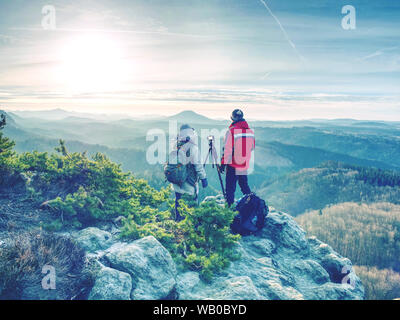 Couple of hikers look down into fogy valley. Photographer stay on cliff and takes photos. Dreamy fogy landscape blue misty sunrise in a beautiful vall Stock Photo