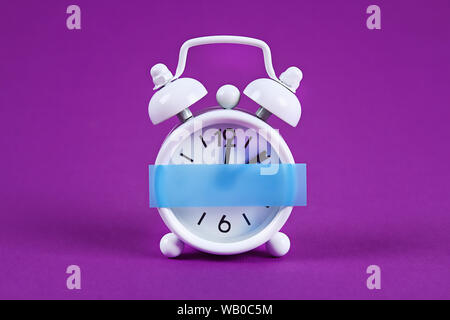 White alarm clock on purple background. Blank sticky note on the alarm clock for text. Reminder. Space for copy. Minimal concept. Stock Photo