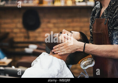 Handsome man with a beard and closed eyes in a black cutting hair cape in the barbershop. Barber in is doing him a face massage. Closeup. Horizontal. Stock Photo