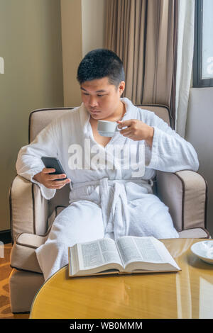Young Asian businessman wearing bathrobe and using smartphone in hotel Stock Photo