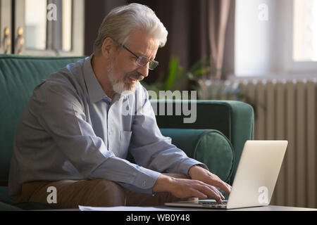Smiling old man in glasses working on laptop at home Stock Photo
