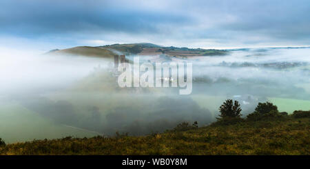 Corfe Castle, Dorset, UK.  23rd August 2019.  UK Weather.  A moody start to the day as the mist begins to break up shortly after sunrise at Corfe Castle in Dorset.  The weather is forecast to be hot and sunny this Bank Holiday weekend.  Picture Credit: Graham Hunt/Alamy Live News